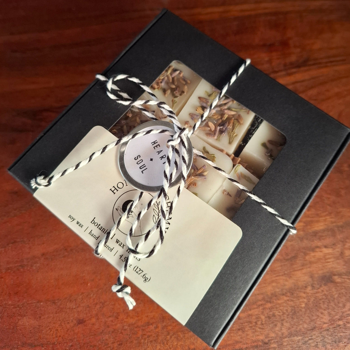 A black gift box containing handmade wax melts, tied with a black and white striped string and a cream paper tag, placed on a wooden surface.