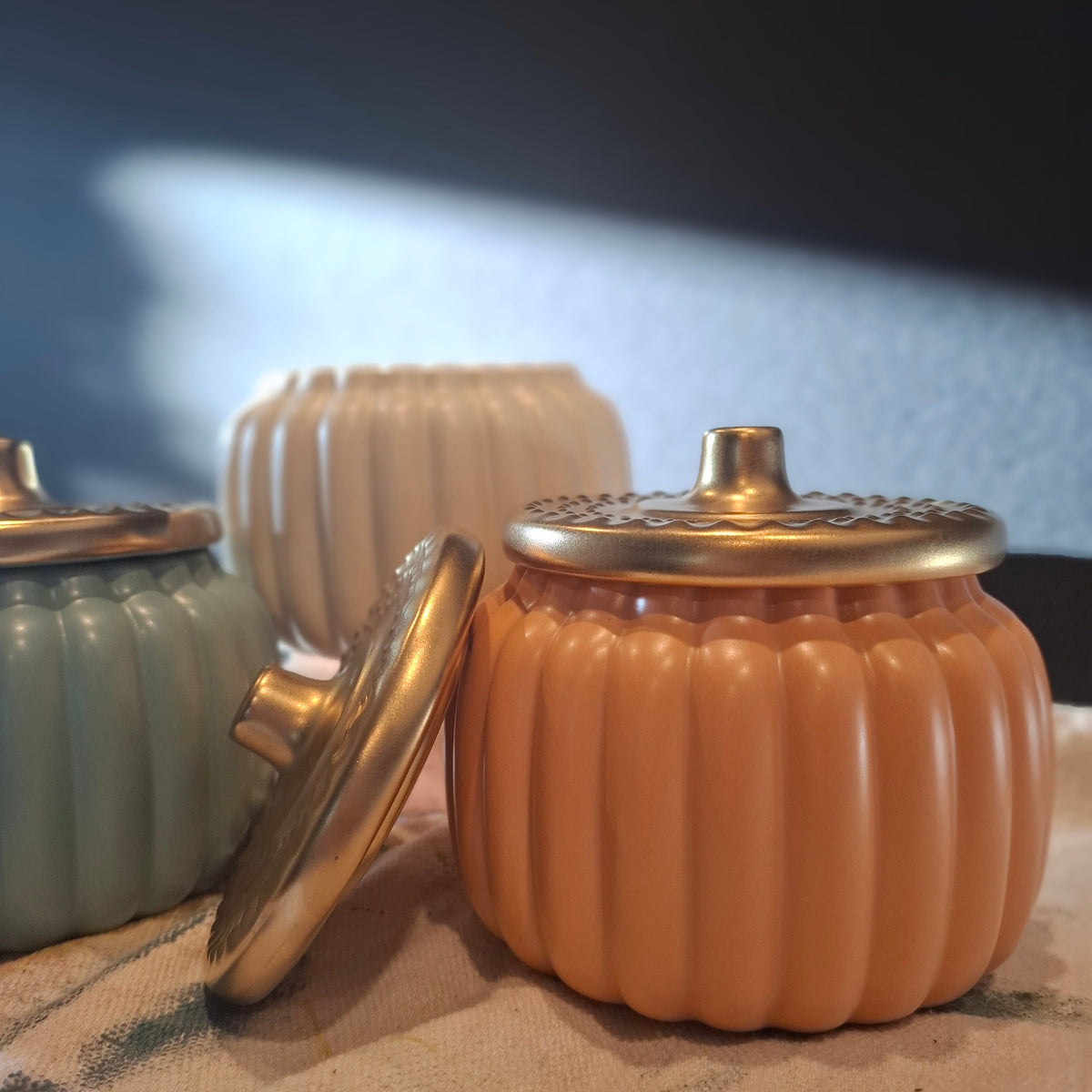 A ribbed decorative jar with a gold-toned lid in a peachy orange color, shown alongside other similar jars in sage green and cream on a textured fabric surface.